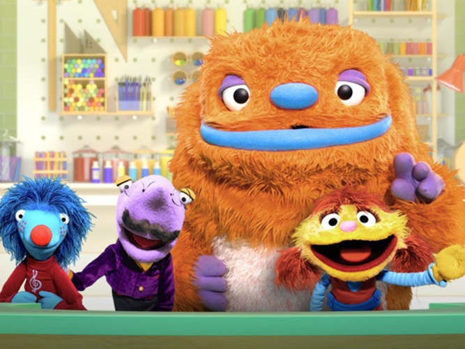 4 friendly monsters wave behind a desk within a colorful workshop.