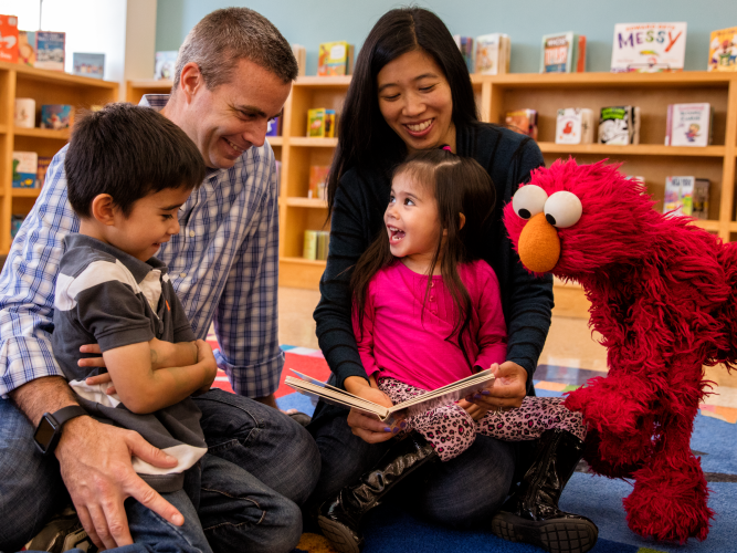 Two parents read to their children with Elmo at a library.