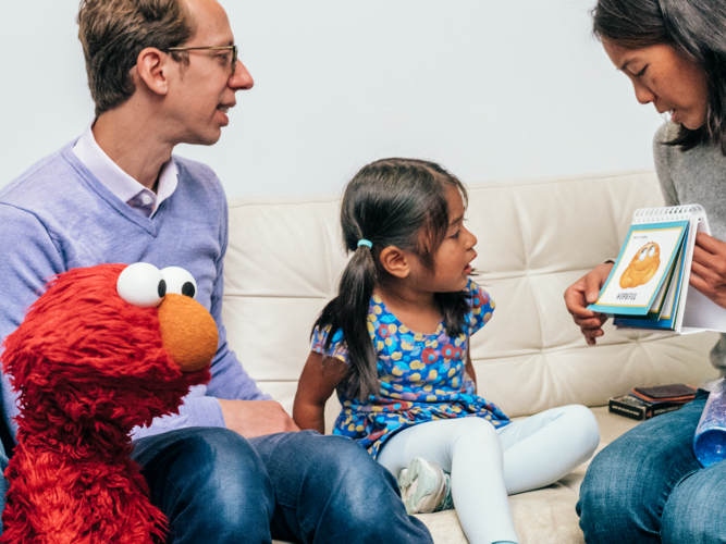 Elmo sitting with a family on a couch as the mother reads aloud.