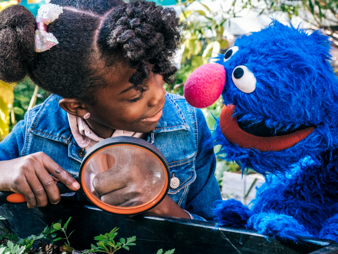 Little girl and Grover looking through a magnifying glass.