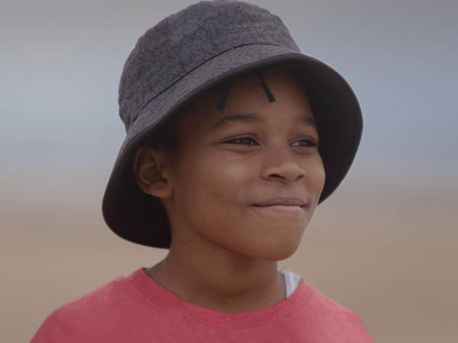 A smiling child stands on a beach wearing a bucket hat and a red shirt
