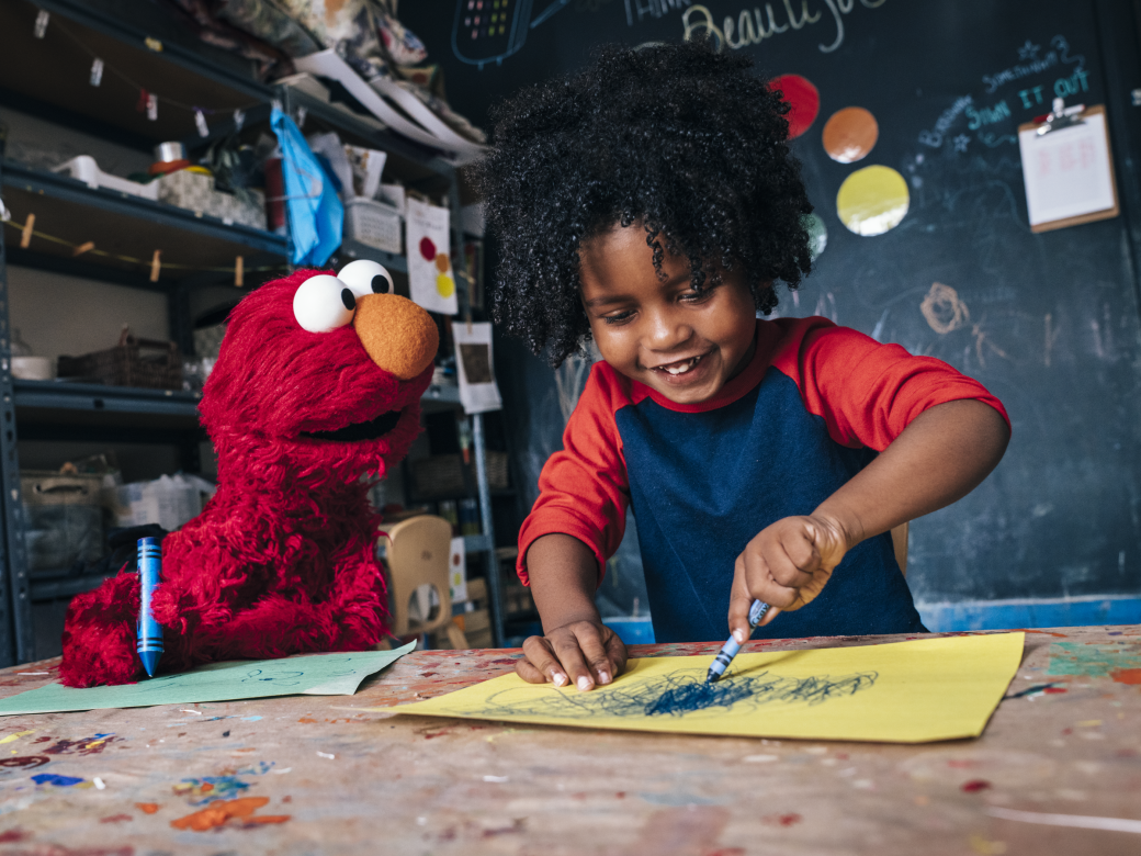 Elmo and a child sit at a table and color together.